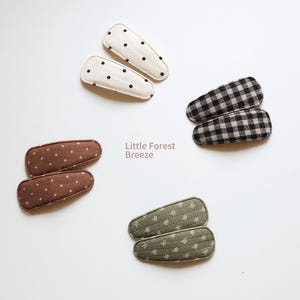 May include: An overhead shot displays four pairs of fabric hair clips in various patterns. Two clips are white with black polka dots, two are brown with white polka dots, two are black and white checkered, and two are green with white dots. The text "Little Forest Breeze" is also visible.