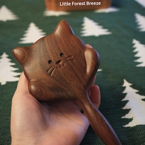 May include: A handcrafted wooden cat-shaped toy with a handle. The toy is made of dark brown wood and features carved details for the eyes, nose, and whiskers. The text "Little Forest Breeze" is visible in the background.