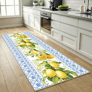 May include: A rectangular kitchen rug with a white center featuring yellow lemons and green leaves. The rug has a blue and white patterned border. The rug is placed on a wooden floor in front of a white kitchen.