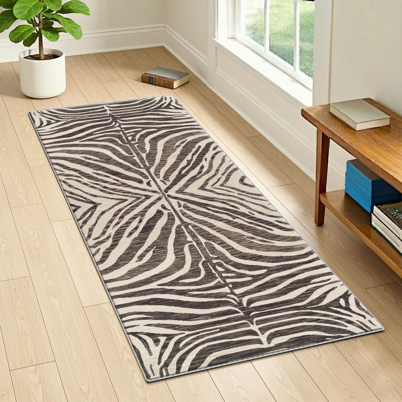 May include: A rectangular rug with a zebra stripe pattern in shades of brown and cream. The rug is placed on a light-colored wooden floor, with a plant and a small wooden table visible in the background. The rug's design is symmetrical.
