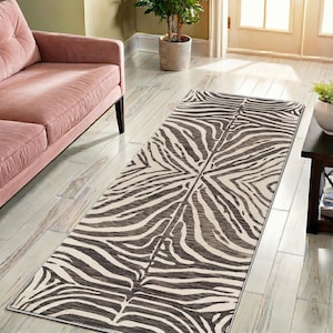Puede incluir: Una alfombra rectangular con estampado de cebra, con fondo crema y rayas marrón oscuro. La alfombra está colocada sobre un suelo de madera clara, cerca de un sofá rosa. El diseño es simétrico, con las rayas convergiendo en el centro.