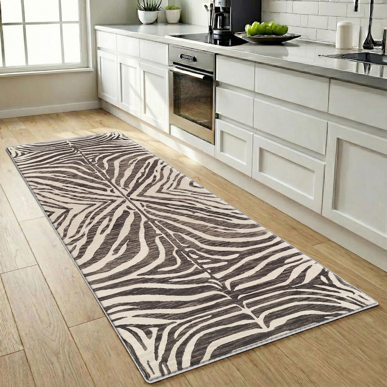 May include: A rectangular zebra-print rug in a kitchen setting. The rug features a pattern of black and cream-colored stripes, creating a bold, animal-inspired design. The rug is placed on a light-colored wooden floor, adding a touch of style to the kitchen.