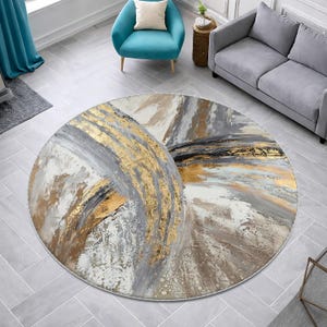 May include: Round area rug with an abstract design. The rug features gold, gray, white, and black brushstrokes. The rug is placed on a gray tiled floor, with a teal chair and gray sofa in the background.