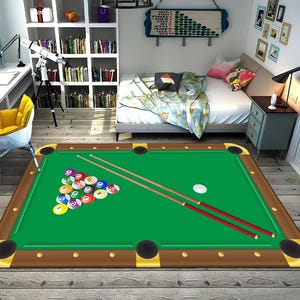 May include: A rectangular rug designed to look like a pool table. The rug has a green playing surface, brown borders, and gold-coloured corner pockets. A rack of billiard balls, two cues, and a white cue ball are visible.