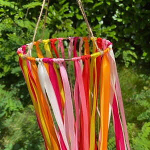 May include: A colourful mobile made of ribbons in shades of pink, orange, and yellow. The ribbons are tied to a metal ring and hang down in a cascade. The mobile is suspended from a string.