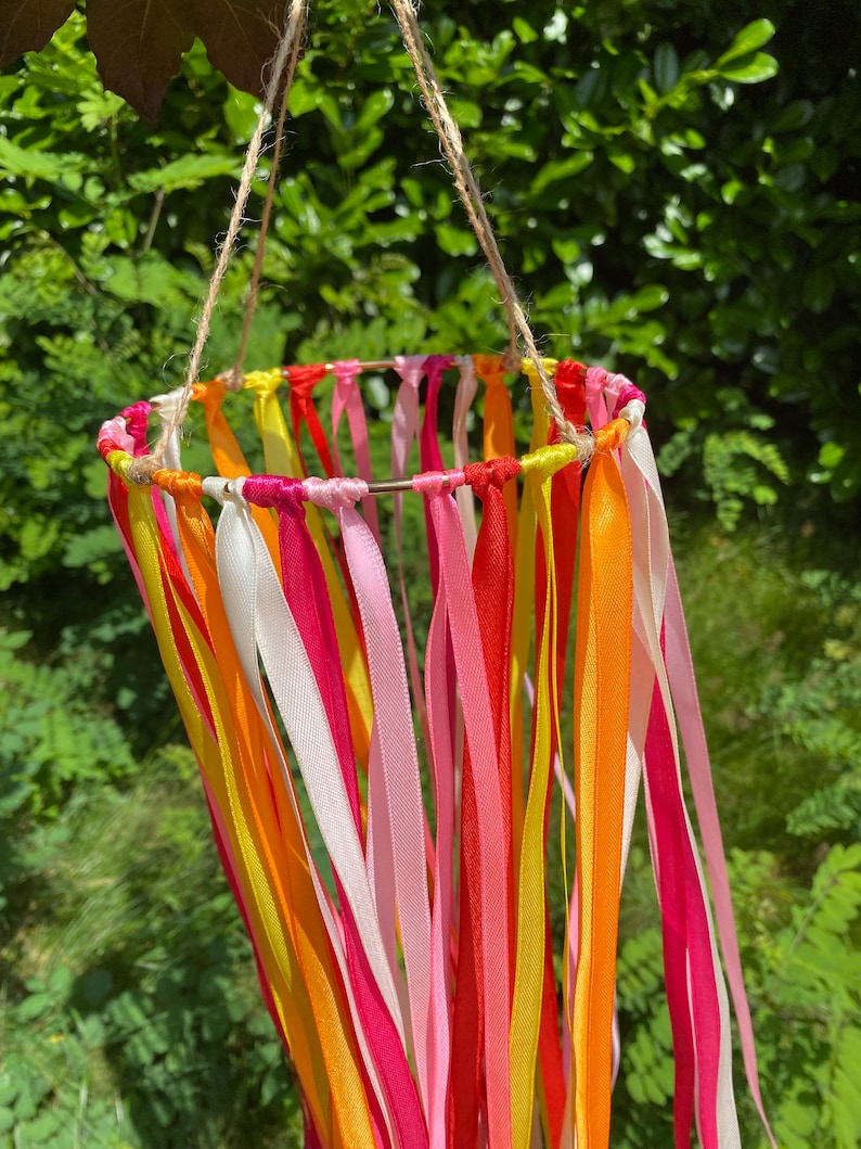 May include: A colourful mobile made of ribbons in shades of pink, orange, and yellow. The ribbons are tied to a metal ring and hang down in a cascade. The mobile is suspended from a string.