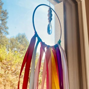 May include: A handmade suncatcher with a metal hoop, wooden beads, and a crystal pendant. Colorful ribbons in red, orange, yellow, green, purple, and pink hang from the hoop. The suncatcher is suspended in front of a window.