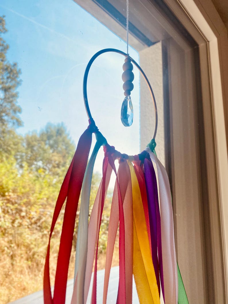 May include: A handmade suncatcher with a metal hoop, wooden beads, and a crystal pendant. Colorful ribbons in red, orange, yellow, green, purple, and pink hang from the hoop. The suncatcher is suspended in front of a window.