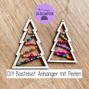 May include: Two wooden Christmas tree ornaments with colorful beads strung across the interior. The trees are light-colored with a triangular shape. A purple circle with text and a rainbow graphic is above the trees. The text "DIY Bastelset Anhänger mit Perlen" is below the trees.