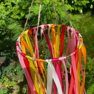 May include: A colourful mobile made of ribbons in shades of pink, yellow, orange, and red. The ribbons are tied to a metal ring and hang down in a cascade.