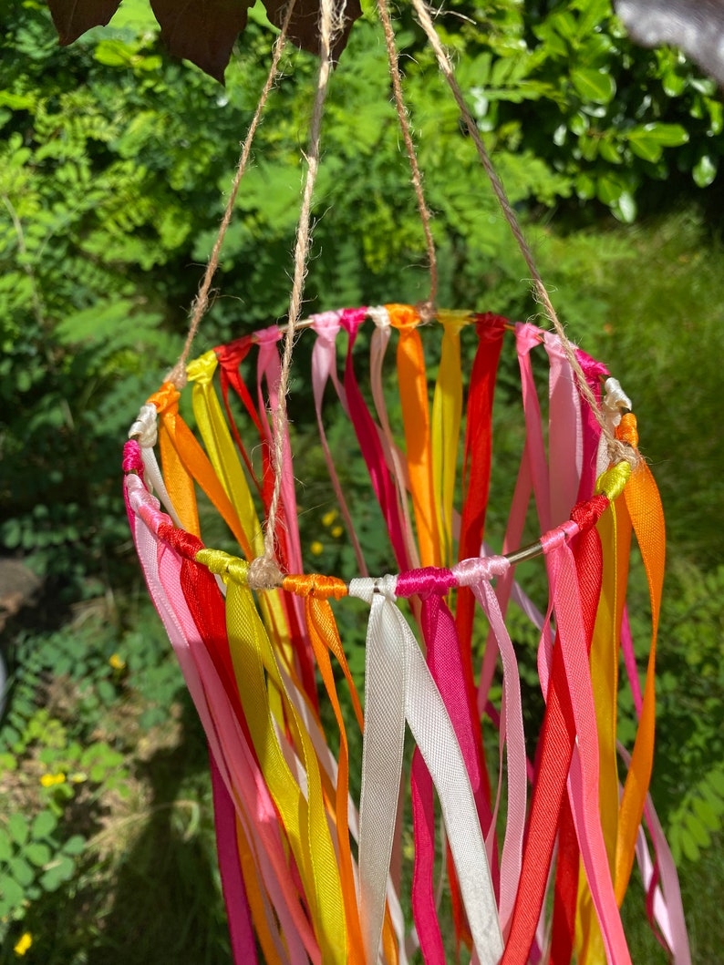 May include: A colourful mobile made of ribbons in shades of pink, yellow, orange, and red. The ribbons are tied to a metal ring and hang down in a cascade.