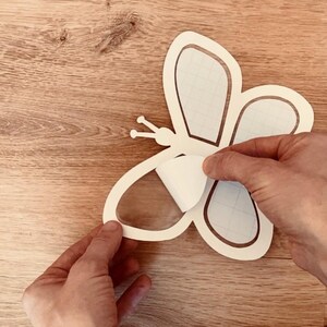May include: A white butterfly-shaped sticker being peeled from a clear transfer sheet. The sticker is on a wooden surface.