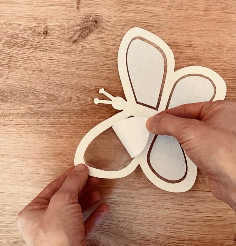 May include: A white butterfly-shaped sticker being peeled from a clear transfer sheet. The sticker is on a wooden surface.