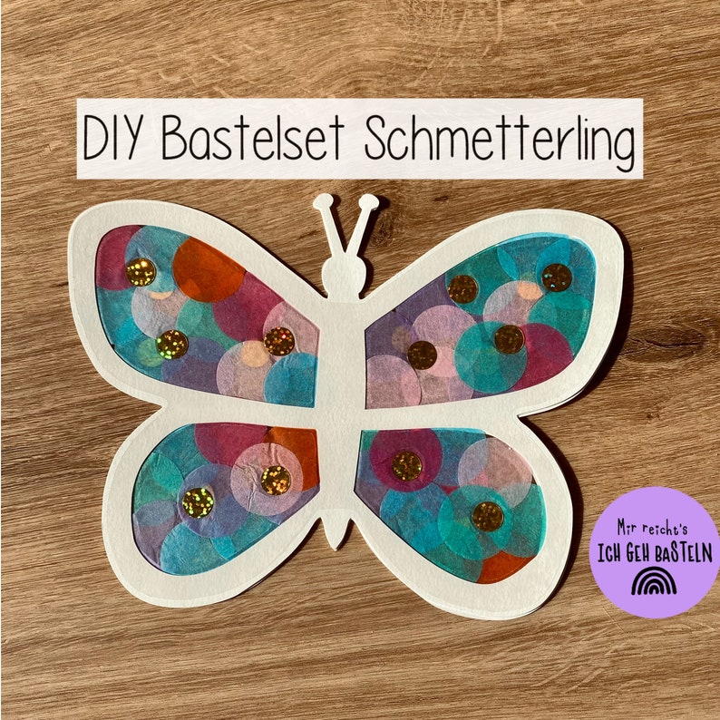 May include: DIY craft kit for making a butterfly. The butterfly is white with a translucent paper design in shades of blue, pink, and purple. The paper design has gold glitter accents. The kit includes a white butterfly cutout and a sheet of translucent paper with a circle design.