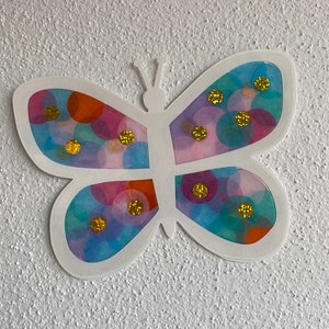 May include: A white paper butterfly with translucent wings decorated with colourful circles and gold glitter.