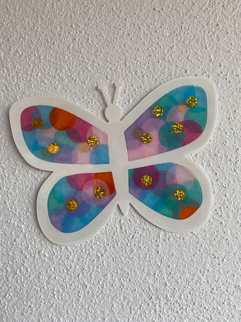 May include: A white paper butterfly with translucent wings decorated with colourful circles and gold glitter.