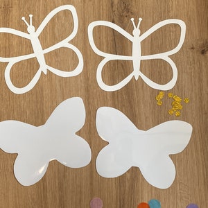 May include: Four white butterfly cut-outs on a wooden surface with colourful confetti and gold glitter. The butterflies are different sizes and shapes.