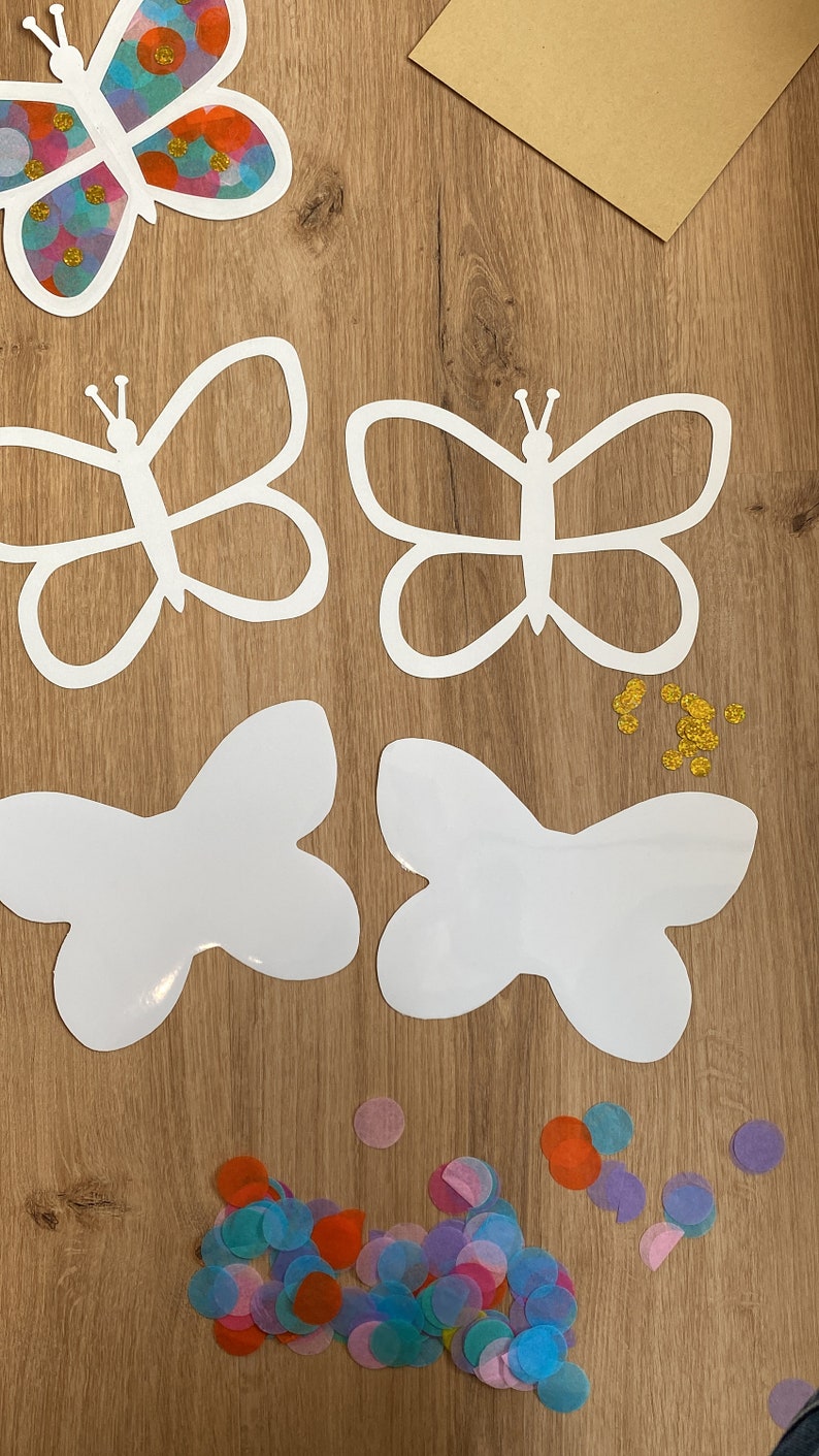 May include: Four white butterfly cut-outs on a wooden surface with colourful confetti and gold glitter. The butterflies are different sizes and shapes.
