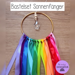 May include: A suncatcher craft kit featuring a gold-colored metal ring with a clear crystal pendant and wooden beads. Rainbow-colored ribbons are attached to the bottom of the ring. The text "Bastelset Sonnenf&auml;nger" is at the top.