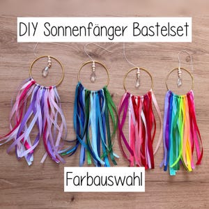 May include: Four DIY suncatcher kits with gold rings, colorful ribbons, and crystal accents. Each kit features a different color scheme: purple, blue, red, and rainbow. Text on the image reads "DIY Sonnenf&auml;nger Bastelset" and "Farbauswahl".