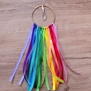 May include: A handmade dream catcher with a gold-colored metal ring. Colorful ribbons in rainbow hues hang from the bottom. A clear crystal and wooden beads are suspended from the top, adding a touch of sparkle and natural elements.