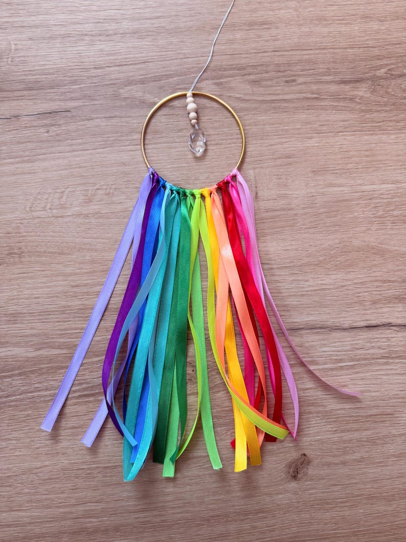 May include: A handmade dream catcher with a gold-colored metal ring. Colorful ribbons in rainbow hues hang from the bottom. A clear crystal and wooden beads are suspended from the top, adding a touch of sparkle and natural elements.