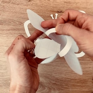 May include: A white paper butterfly craft with a cut-out design. The butterfly is being folded in half to create a three-dimensional shape.