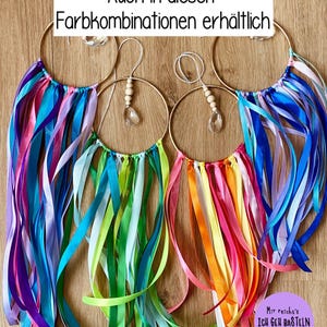May include: Four decorative hoop ornaments with colorful ribbons in shades of blue, pink, green, and orange. Each hoop has a gold-colored ring and a crystal or wooden bead accent. The text "Auch in diesen Farbkombinationen erh&auml;ltlich" is visible.