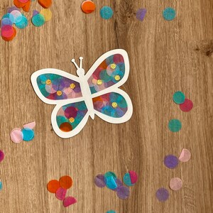 May include: A white butterfly cutout with colourful tissue paper circles glued inside. The butterfly is on a wooden surface with more tissue paper circles scattered around it.