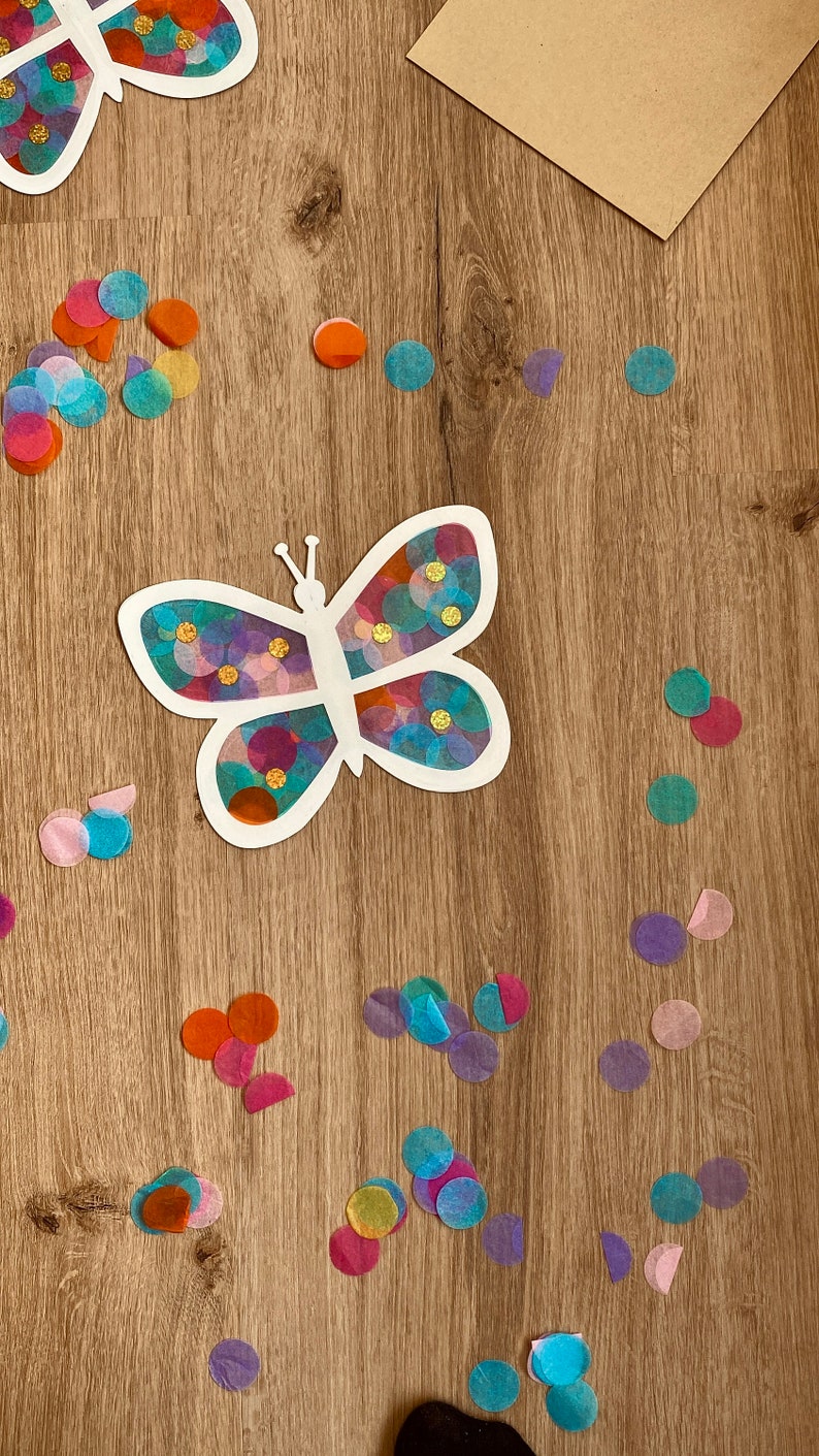 May include: A white butterfly cutout with colourful tissue paper circles glued inside. The butterfly is on a wooden surface with more tissue paper circles scattered around it.