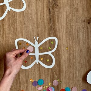 May include: A white paper butterfly cutout with a pink, blue and orange confetti design. The butterfly is being decorated with colourful tissue paper circles.