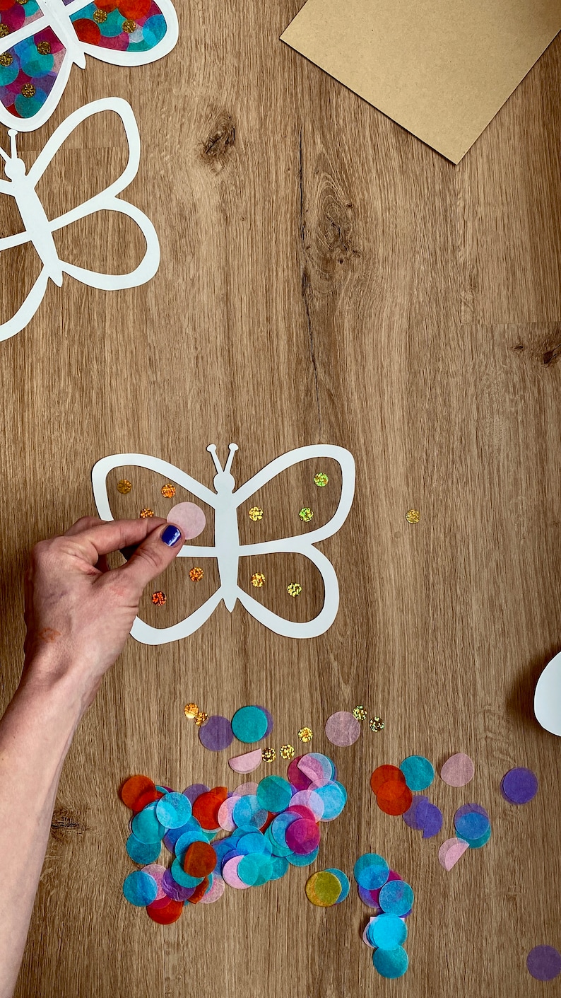 May include: A white paper butterfly cutout with a pink, blue and orange confetti design. The butterfly is being decorated with colourful tissue paper circles.