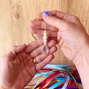 May include: A clear crystal pendant with wooden beads suspended from a string, held in hands with purple nail polish. A gold ring and colorful ribbons are visible. The pendant is teardrop-shaped.