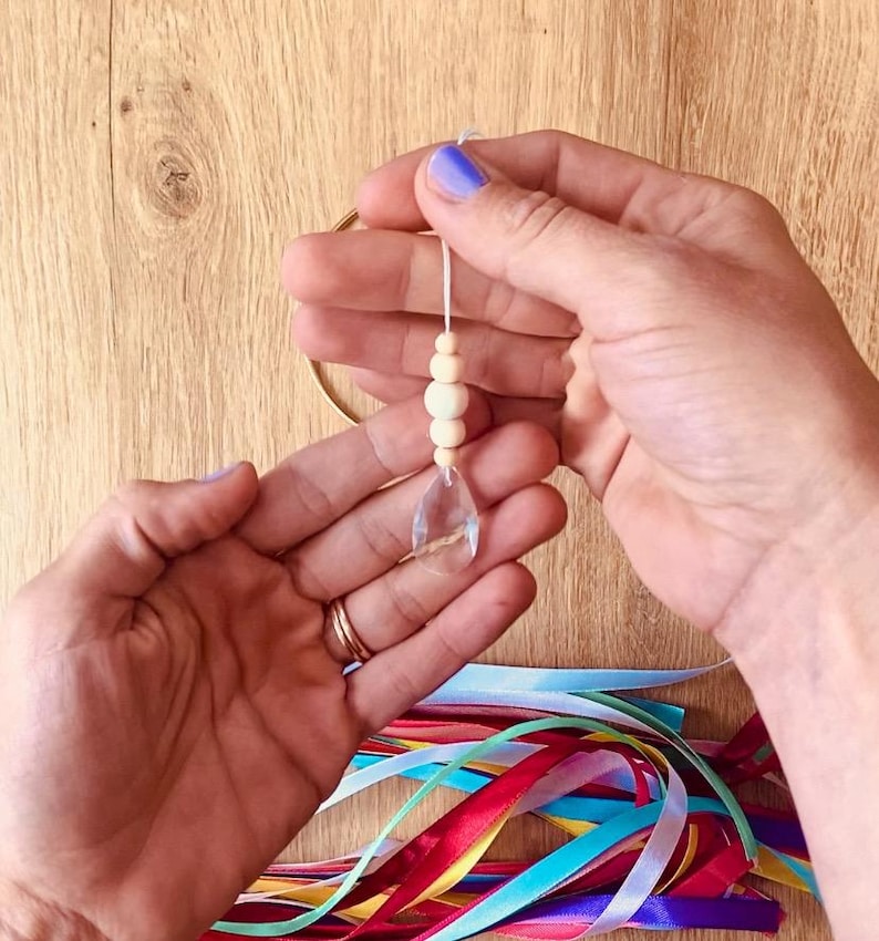 May include: A clear crystal pendant with wooden beads suspended from a string, held in hands with purple nail polish. A gold ring and colorful ribbons are visible. The pendant is teardrop-shaped.
