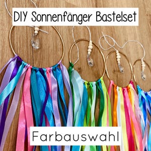 May include: A collection of colorful DIY suncatcher kits. Each kit features a gold metal hoop adorned with vibrant ribbons in shades of pink, purple, blue, and green. Clear crystal-like beads and wooden beads dangle from the top of each hoop. The text "DIY Suncatcher Kit" and "Color Selection" are visible.
