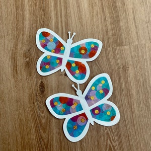 May include: Two white paper butterflies with colourful circle patterns. The butterflies have gold glitter accents.