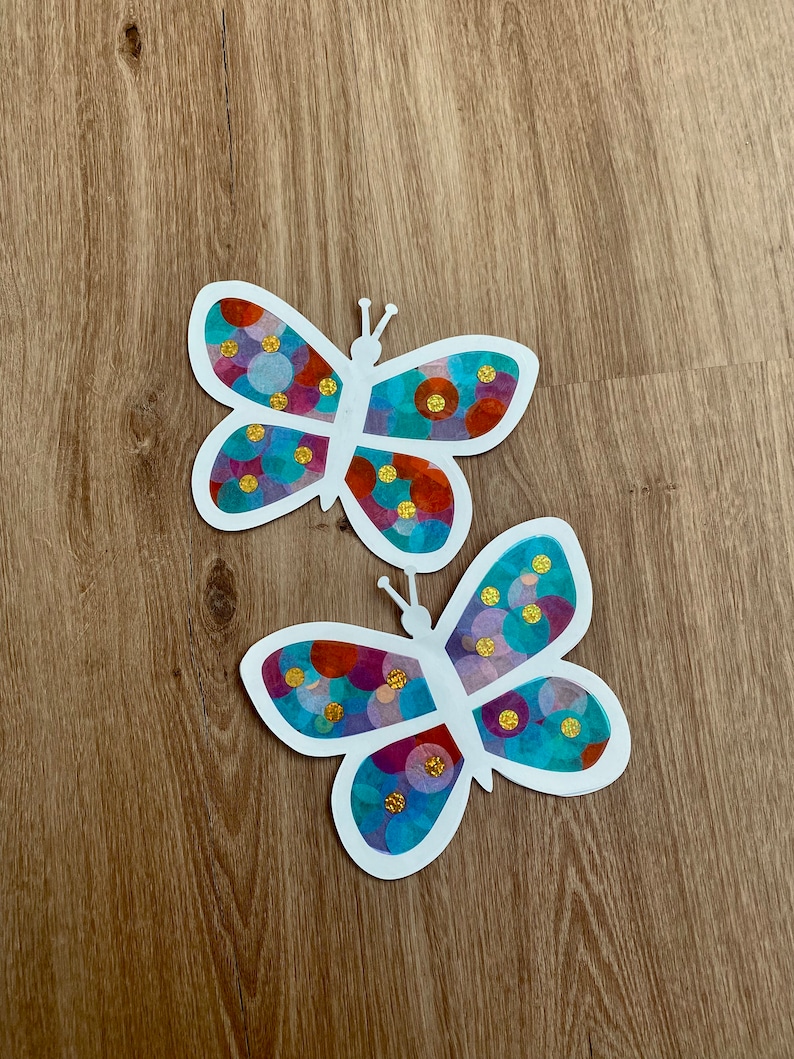 May include: Two white paper butterflies with colourful circle patterns. The butterflies have gold glitter accents.