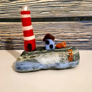 May include: A handcrafted felted wool sculpture depicting a coastal scene. It features a red and white striped lighthouse, a small blue house, and an orange buoy on a stone base. An orange ladder is on the side of the stone.