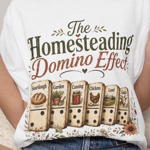 May include: A white short-sleeve t-shirt featuring the text The Homesteading Domino Effect in dark green and brown. Below the text are six dominoes labeled Sourdough, Garden, Canning, Chickens, Land, and Goats, each with a corresponding illustration.