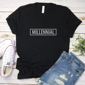 May include: Black v-neck t-shirt with the word "MILLENNIAL" printed in white on the front. The shirt is paired with denim shorts and white sneakers.