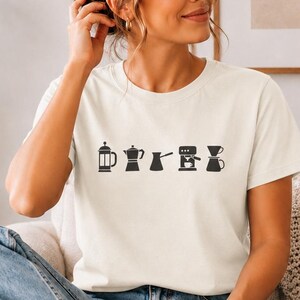 May include: A cream-colored short-sleeve t-shirt featuring a horizontal row of black graphic icons depicting various coffee brewing methods, including a French press, a moka pot, a cezve, an espresso machine, and a pour-over coffee dripper.
