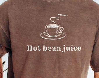 Könnte beinhalten: Ein braunes Kurzarm-T-Shirt mit einer weißen Strichzeichnung einer Kaffeetasse auf einer Untertasse, aus der Dampf aufsteigt. Unter der Grafik ist der Text "Hot bean juice" in einer einfachen, weißen, serifenlosen Schrift auf die Brust gedruckt.