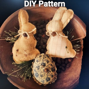 May include: Two brown fabric bunnies and a floral fabric egg in a wooden bowl. The bunnies have bells on their chests and are tied with twine. The egg has a button on the top. DIY Pattern.