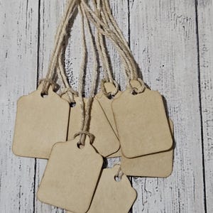May include: A collection of blank, light brown wooden tags with rounded corners and a hole at the top for string. Each tag is attached to a natural twine string. The tags are arranged against a weathered, white-painted wooden background.