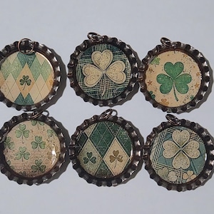 May include: Six circular pendants with a shamrock theme. Each pendant features a different design, including shamrocks and diamond patterns in green, white, and beige, set within a metal bottle cap frame. Each has a small loop for hanging.