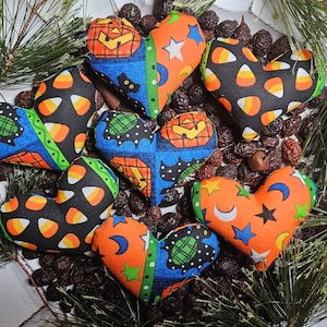 May include: Six stuffed fabric hearts featuring various Halloween-themed patterns including orange jack-o'-lanterns, black candy corn, blue backgrounds with stars and moons, and green accents. The hearts are arranged in a cluster on a dark surface.