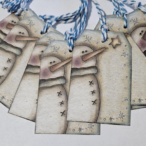 May include: Set of six rustic snowman gift tags with blue and white twine. Each tag features a hand-painted snowman with a brown nose and black eyes. The tags are decorated with snowflakes and a star.