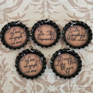 Könnte beinhalten: Fünf schwarze Flaschenverschluss-Anhänger mit verschiedenen inspirierenden Zitaten darauf gedruckt. Die Zitate lauten: "Live Laugh Love", "Do The Impossible", "Do Everything With Love", "Live Laugh Love" und "Dance Like You Mean It".