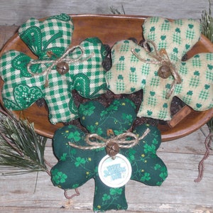 Puede incluir: Cuatro tréboles de tela en un cuenco de madera, decorados para el Día de San Patricio. Los tréboles presentan patrones verdes y blancos, atados con hilo y pequeñas campanas. Un trébol tiene una etiqueta que dice "Happy St. Patrick's Day".