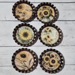 Könnte beinhalten: Sechs dekorative Flaschenverschluss-Magnete mit Sonnenblumenmotiven. Jeder Verschluss zeigt eine andere Sonnenblumenanordnung mit gelben und braunen Farbtönen. Die Verschlüsse haben einen metallischen, strukturierten Rand und sind auf einer weißen Holzoberfläche angeordnet.